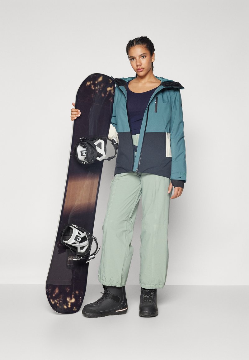 Snowboarder hält ein dunkles Snowboard mit weißen Bindungen, trägt eine türkisfarbene und schwarze Jacke, grüne Hose und schwarze Stiefel. Glatte Texturen.