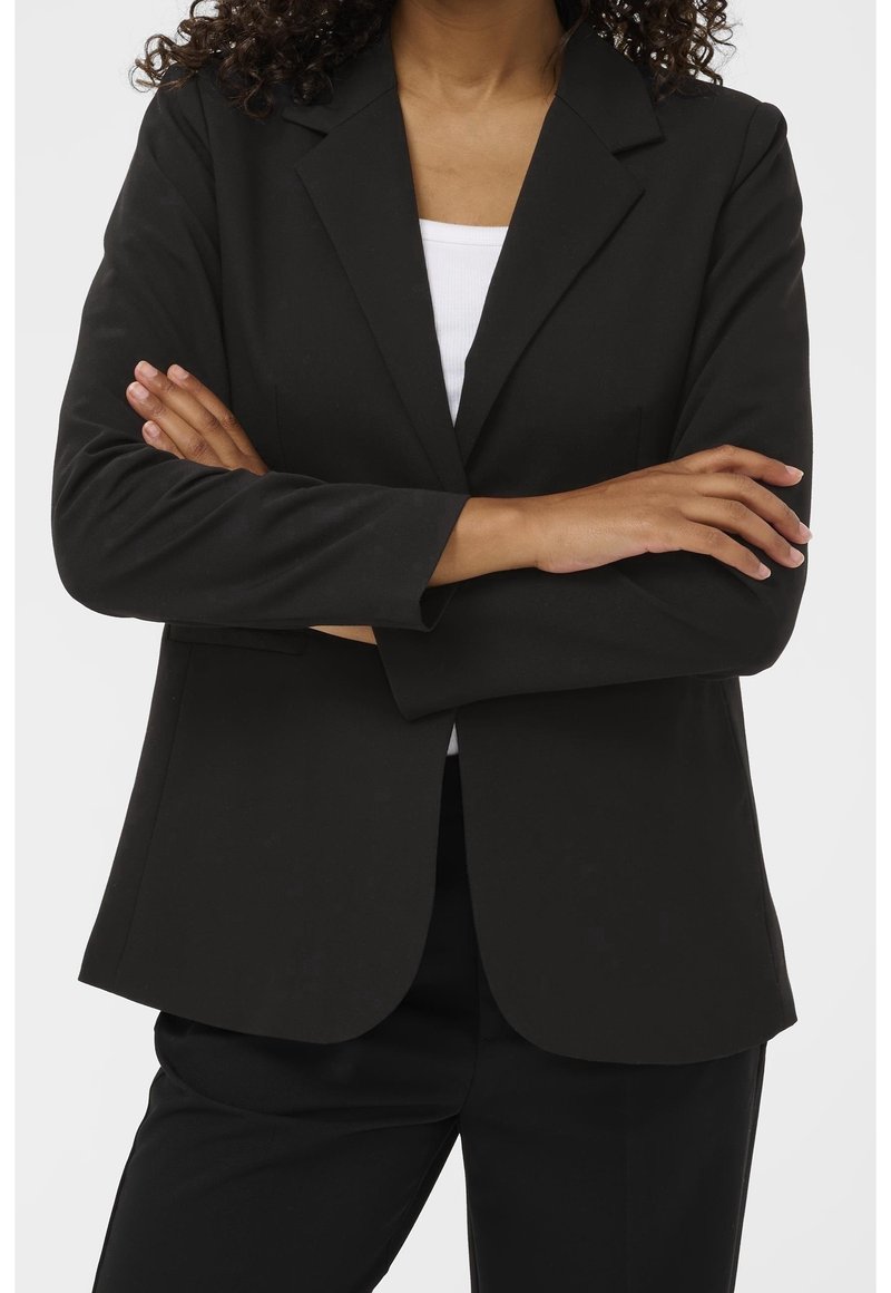 Femme aux cheveux bouclés portant un blazer noir et un pantalon noir, les bras croisés, avec un haut blanc visible sous le blazer.
