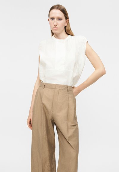 Γυναίκα με ίσια ανοιχτοκάστανα μαλλιά φοράει λευκό αμάνικο πουκάμισο με γεμισμένους ώμους και χαλαρά μπεζ παντελόνια ψηλόμεσα, με το χέρι στη μέση.