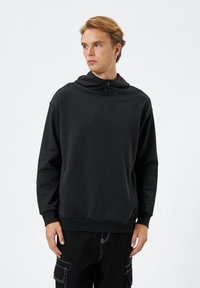 Svart huvtröja i mjukt tyg, med hög krage och knappdetalj, känguruficka och ribbade ärmslut.