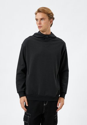 Sudadera negra con capucha hecha de una tela suave, que presenta un cuello alto con detalles de botón, bolsillo tipo canguro y puños de canalé.
