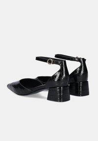 Zapatos negros de charol con tacón cuadrado, punta afilada, correa en el tobillo y textura de cocodrilo. Presentan una hebilla dorada.