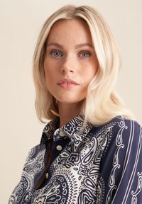 Blusa a righe blu navy e bianca con colletto, dotata di bottoni decorativi e tessuto fluido con disegni intricati.