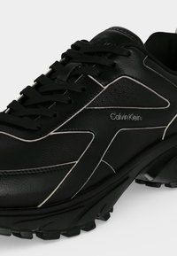 Μαύρο δερμάτινο αθλητικό παπούτσι Calvin Klein με μαύρα κορδόνια, λευκές ραφές και ανθεκτική, υφή σόλα σε γκρι φόντο.