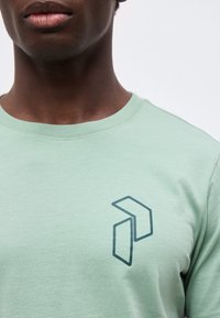 Κοντινό πλάνο ενός ατόμου που φοράει ανοιχτοπράσινο μπλουζάκι με σκούρο γεωμετρικό λογότυπο στο αριστερό στήθος, ορατό από τον λαιμό μέχρι το πάνω μέρος του κορμού.