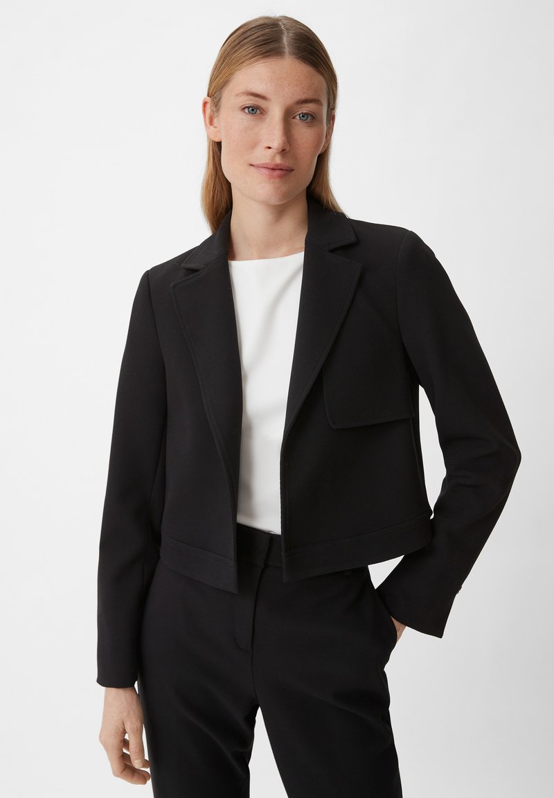 Zwarte cropped jas van gestructureerde stof met een revers en een patchzak; gecombineerd met een witte top en zwarte broek.
