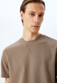 Brun tekstureret bomulds t-shirt med rund hals, der har et dekorativt stikmønster i form af et 'X' ved halsudskæringen.