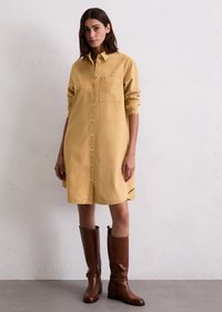 Vrouw in een mosterdkleurige button-up shirtjurk met lange mouwen en bruine leren laarzen tot de knie, staande tegen een witte textuurmuur.