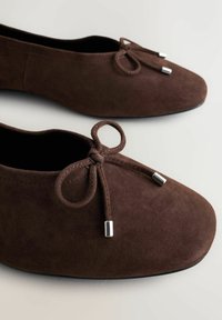 Barna szarvasbőr balettcipők lekerekített orral és díszes masnival, amely ezüst fém végekkel van ellátva, világos háttéren.