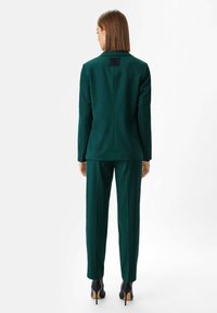 Green tailored suit with a fitted blazer and straight-leg trousers. Features a textured fabric and a small logo patch at the back of the blazer.