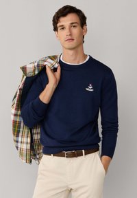 Homme aux cheveux bruns courts portant un pull marine et un pantalon beige, tenant une veste à carreaux multicolores sur l'épaule, sur un fond uni.