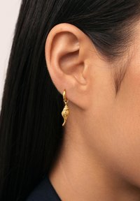 Pendiente en forma de concha dorada que cuelga de un aro, con superficie texturizada visible que muestra un diseño detallado. Se lleva en un primer plano de la oreja.