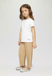 Kind met een wit katoenen T-shirt met een klein zwart logo, beige geplooide broek en metallic zilveren schoenen met strikken.