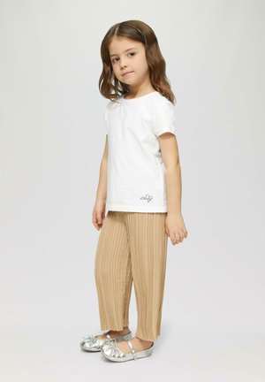 Kind, das ein weißes Baumwoll-T-Shirt mit einem kleinen schwarzen Logo, beigefarbenen Plisseehosen und metallisch silbernen Schuhen mit Schleifen trägt.