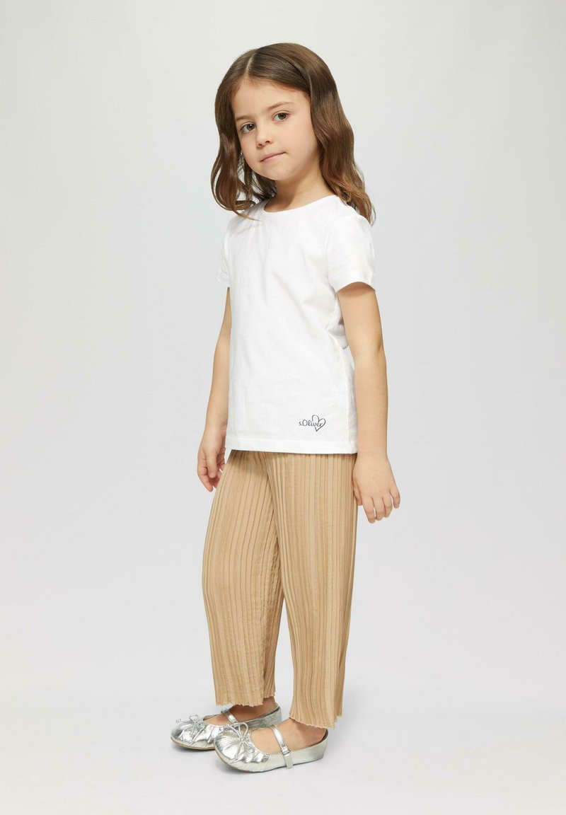 Kind met een wit katoenen T-shirt met een klein zwart logo, beige geplooide broek en metallic zilveren schoenen met strikken.