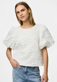 Crop top bianco con tessuto testurizzato e maniche a sbuffo, dotato di scollatura rotonda e un motivo di tessuto increspato. Abbinato a pantaloni corti di jeans azzurri.