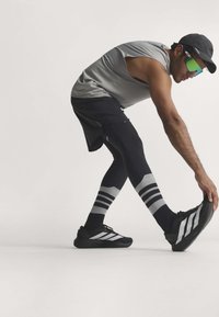 Uomo con cappello e occhiali da sole che allunga una gamba in avanti toccando il piede, vestito con canotta grigia, pantaloncini neri, leggings e scarpe da ginnastica.