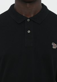 Tricou polo negru din țesătură texturată, având un guler cu trei butoane și un mic logo brodat cu un zebra multicolor pe piept.