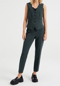 Gilet ajusté vert foncé avec cinq boutons noirs, associé à un pantalon slim assorti et des mocassins noirs imposants, avec une texture lisse.