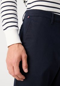 Mörkblå bomullshängselbyxor med en slät yta, prydda med en liten röd, vit och blå etikett vid midjan. Hand som vilar på sidan.