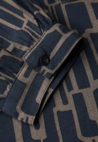 Tissu bleu foncé avec un motif géométrique beige, présentant un bouton et un détail de poignet plié. Texture lisse et design structuré.