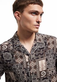 Junger Mann mit kurzen braunen Haaren, der ein braun-schwarz gemustertes Hemd mit Knöpfen trägt und nach rechts vor einem weißen Hintergrund schaut.