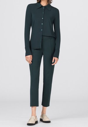 Kobieta w ciemnozielonej koszuli zapinanej na guziki i dopasowanych spodniach o długości 7/8, w beżowych lordach, stoi na tle jasnego, gładkiego tła.