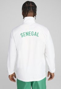 Hvid jakke med høj krave, prydet med "SENEGAL" i grønne bogstaver på ryggen. Stoffet ser glat og let ud.