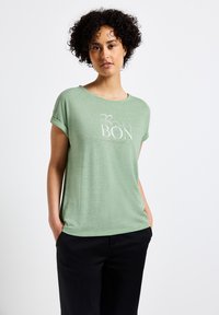Zelené tričko s krátkým ruk�ávem a kulatým výstřihem, s ohrnutými manžetami, se bílým potiskem textu "BON" a měkkou, texturovanou látkou.