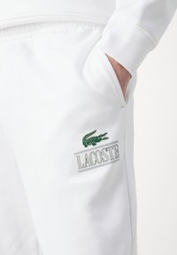 Bílé bavlněné tepláky se bočními kapsami, s zeleným logem krokodýla Lacoste a horizontálním nápisem značky. Měkká struktura a volný střih.