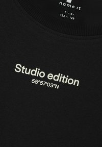 Černý bavlněný svetr s kulatým výstřihem, na němž je bílé písmo: "Studio edition 55°57'03"N." Měkká textura, volný design.