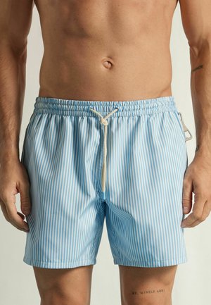 Hombre con pantalones de baño a rayas verticales azules y blancas con cordón, de pie frente a un fondo claro y liso.