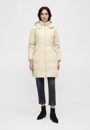 Donna che indossa un cappotto imbottito beige chiaro con cappuccio, pantaloni neri sopra la caviglia e stivaletti neri, in piedi davanti a uno sfondo semplice.