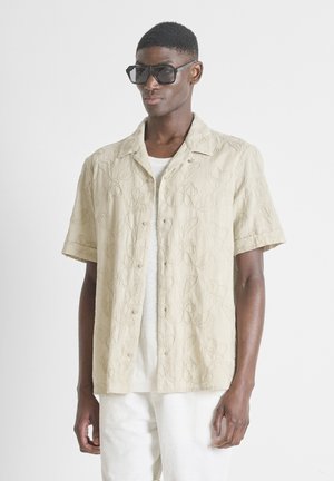 Muškarac nosi prevelike crne kvadratne sunčane naočale i bež košulju kratkih rukava s diskretnim uzorkom lišća.