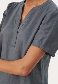 Blouse à manches courtes et col en V, de couleur gris ardoise, avec un tissu texturé et de petites manches bouffantes, conçue avec une coupe décontractée.