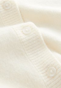 Mīksts krēmkrāsas džempera audums ar ribotu tekstūru un gludu plāksteri. Aizdares arapaļi balti pogas.