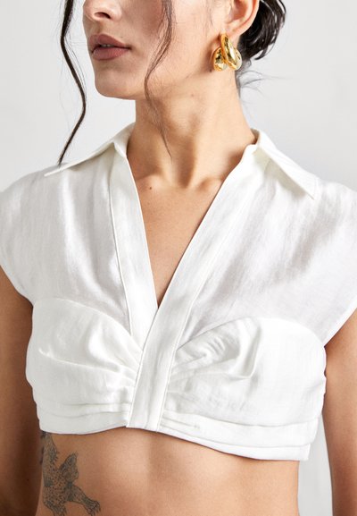 Top court blanc à manches courtes, avec un détail plissé à l'avant et un col en V. Textile lisse et tissu léger. Boucles d'oreilles dorées visibles.