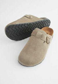 Sabots en suede beige avec un dos ouvert et une sangle sur le dessus, dotés d'une semelle en caoutchouc noir texturé avec un motif en zigzag.
