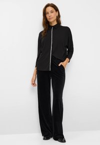 Frau steht mit der Hand in der Tasche, trägt ein schwarzes Hemd mit Strassdetails und schwarze weit geschnittene Hosen vor weißem Hintergrund.