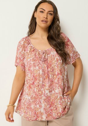 Kvinde med langt brunt hår iført en løs lyserød og orange blomsterbluse med korte ærmer og beige bukser, stående mod en neutral baggrund.