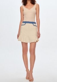 Top a canotta beige a coste con scollatura a V profonda, abbinato a una gonna crema testurizzata con orlo a balze e dettagli blu su tasche e cintura.