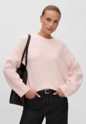 Femme portant un pull rose clair, un jean foncé, de petites boucles d'oreilles en forme d'anneau, les mains dans les poches et portant un sac à bandoulière foncé.