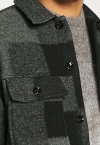 Chaqueta de lana de cuadros gris oscuro y negro con botones negros sobre un suéter borroso gris claro, mostrando detalles del cuello y la parte superior del pecho.