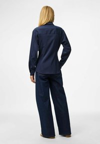 Person with long blonde hair wearing a navy blue pinstripe shirt and wide-leg dark blue jeans, standing with one hand on hip, facing away.