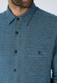 Nærbillede af en mand iført en struktureret teal-farvet skjorte med sorte knapper og krave, hvor man kan se dele af hans skæg og hals.