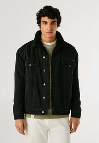 Chaqueta de mezclilla negra con un suave cuello forrado de piel, bolsillos frontales y botones plateados, llevada sobre un suéter a rayas verde.