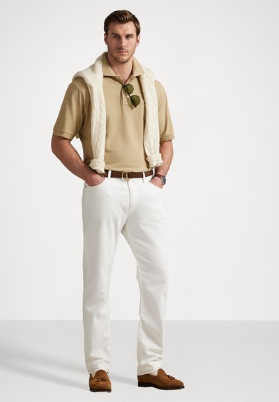Camisa polo bege combinada com calças brancas, um suéter creme de ponto canelado colocado sobre os ombros, e sapatos moccasins castanhos.