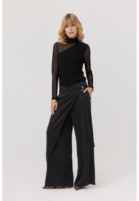 Kobieta w czarnej bluzce z przezroczystymi rękawami, szerokich spodniach w fasonie wrap oraz butach na obcasie w panterkę, stojąca z ręką w kieszeni na jednolitym tle.