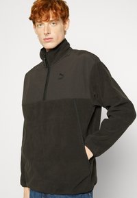 Svart fleece genser med høy krage, glidelås foran og teksturerte seksjoner. Har en liten logo på brystet og sidelommer.