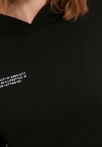 Svart hoodie med en mjuk textur, med huva och en vit textgrafik på framsidan som säger: "Skönhet i enkelhet som livsstil är att släppa taget."